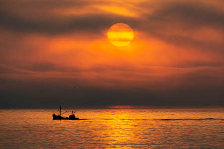 fishing boat transiting the sea in contrast to orange sunsetの写真素材