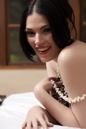 Sexy naked young caucasian adult woman with red lips, short black hair and a pierced eyebrow, covered in a dark satin sheet and lying on a bed, wearing a string of white pearlsの写真素材