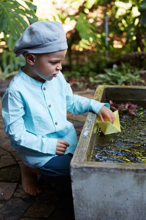 Little four year old boy in a blue outfit playing with paper boats in a small outdoor water fountainの写真素材