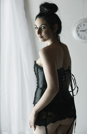 Beautiful, voluptuous and sexy caucasian adult woman in black lace up corset and garters in a boudoir setting lit from the side with window light の写真素材