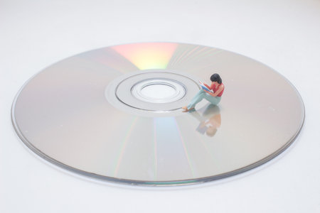 A miniature figure sits on a shiny compact disc, reading a book.