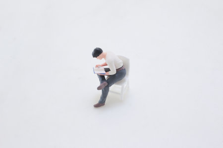 A tiny figurine sits on a white chair, absorbed in a bookの写真素材