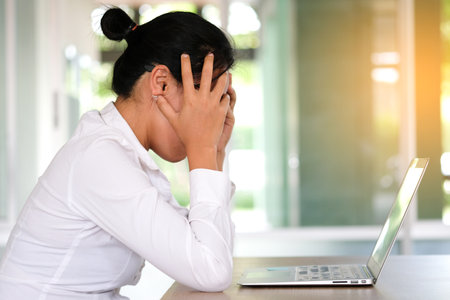 woman sitting down, his face unsettled. At the computer desk she has headaches and stress. Cause of hard work and insufficient rest.の写真素材