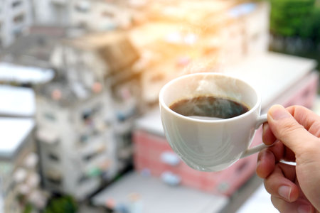Hand of a man holding a hot coffee cup with smoke at the new morning on a modern city backgroundの写真素材