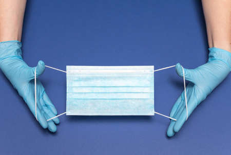 Hands in blue medical gloves is holding face mask on blue background. Protective respiratory medical mask against spreading virus. Close up, copy space, top view, flat lay. High quality photoの写真素材