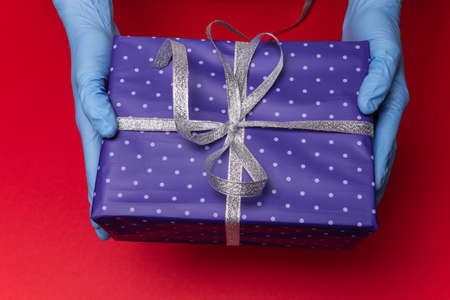 Hands in blue latex gloves holds blue gift box in paper with polka dots with tied ribbon with as a present for Christmas, new year, valentine day or anniversary on red background. High quality photoの写真素材