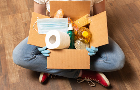A volunteer has sitting on the floor and holding donation box with food, toilet paper and sanitizer. Food delivery service for those who need. High quality photo.の写真素材
