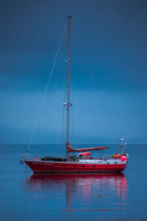 Anchored sailboat reflecting on the sea water on a cloudy day and blue background in Ushuaia, Tierra...