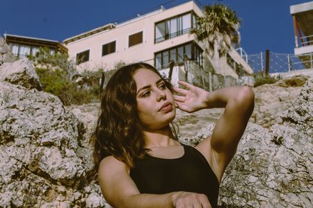 Young latin woman proud of her strong body. Fit girl showing her strong arms in the water. concept of being proud of your body, and displaying it on the beach. Woman in black swimsuit on the rocks.の写真素材