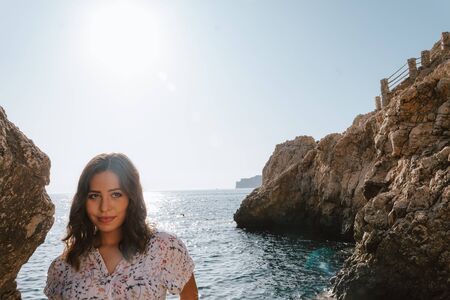 Adventurous young latin woman looking sideways on the sea rocks. Woman on cliff, with the sunset. Jovial near the sea on vacation in the Mediterranean with turquoise blue water.の写真素材