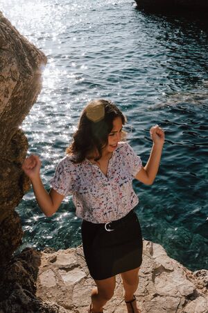 Happy young woman dancing by the sea, celebrating her vacation or the beginning of the school season in her new destination. Woman on cliff, with the sunset. Jovial near the sea on vacation in the Mediterranean with turquoise blue water.の写真素材