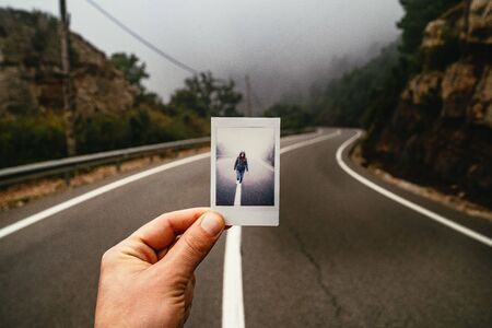 Man holding a photo of a person walking on the centre white line in a misty road lining it up with the misty landscape in front of him in the Monestir de Monserrat Region, Barcelona, Spainの写真素材
