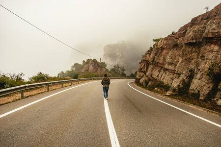 Person walking along a centre line in a road winding through the mountains in the Monestir de Montserrat Region near Barcelona Spain on a cold misty dayの写真素材