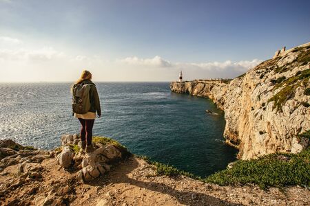 Female hiker admiring view while standing on edge of rocky cliff covered in small plants next to body of waterの写真素材
