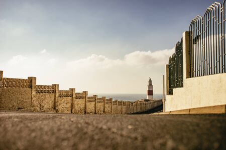Worms eye view of short decorative walls with black fencing bordering asphalt road to lighthouse in distanceの写真素材