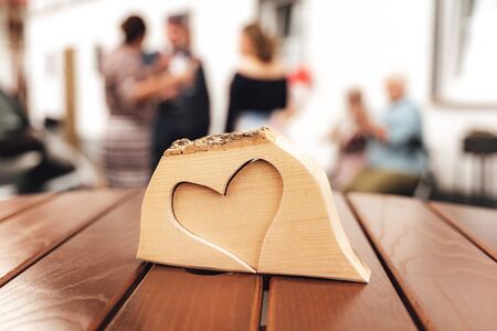 Heart shape carved in slice of wood laying on wooden table. Symbol of love. Blurred people in background.の写真素材