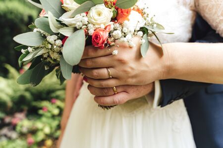 Closeup view of newlyweds hands holding colorful wedding bouquet. Bride and groom wearing wedding rings. Outdoor background. Wedding day concept.の写真素材