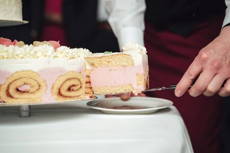 Wedding cake standing on festive table. Closeup on male hands serving piece of cake at white plate....