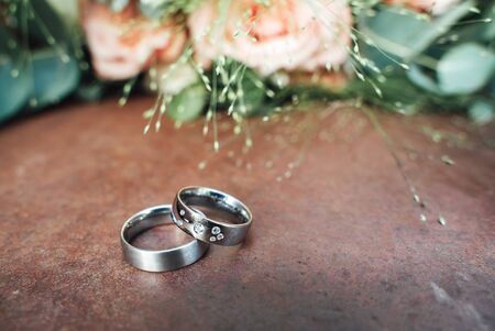 Close up of two wedding rings laying on brown color stone surface. Blurred wedding bouquet in background. Wedding day concept.の写真素材