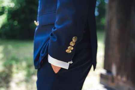 Side view of man in blue suit with hand in pocket. Close up of jacket sleeve gold color buttons with anchor sign. Outdoor background.の写真素材