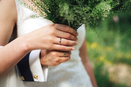 Closeup view of newlyweds hands holding colorful wedding bouquet. Bride and groom wearing wedding rings. Outdoor background. Wedding day concept.の写真素材