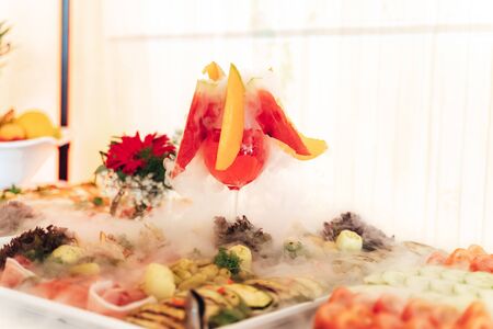 Smoking glass at fruit and vegetable buffet table. Closeup of glass of red beverage with white dense fog. Chemical reaction of dry ice with water.の写真素材