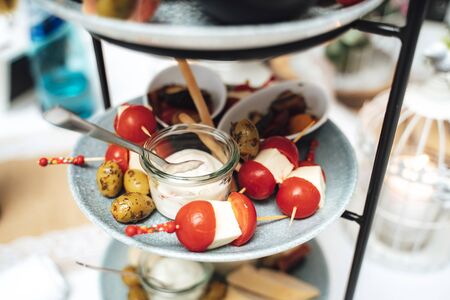 Multi-tiered plate stand with small snacks and ingredients. Stuffed cherry tomatoes, olives and glass of white dip. Celebration, party, weddingの写真素材
