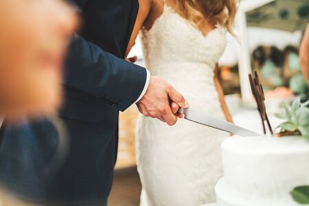 Close up of wedding cake cutting. Newlyweds holding knife together and slicing white wedding cake. Wedding day concept.の写真素材