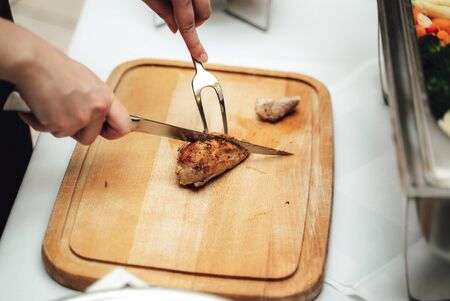 Man preparing dish. Male hands slicing piece of roasted pork meat with knife and fork on wooden board. Celebration, party, birthday or weddingの写真素材