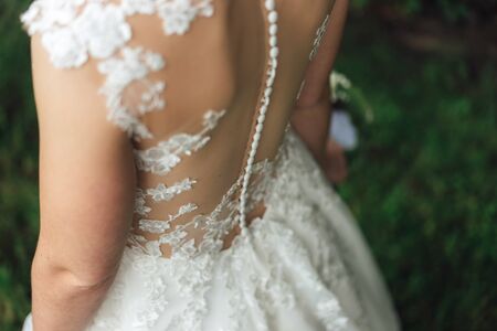 Bride in wedding dress. Close up of lace decorated back of white wedding gown and dense row of white buttons on spine. Outdoor background. Wedding dayの写真素材