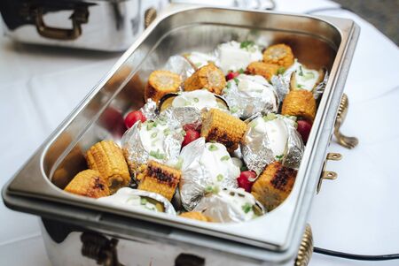 Grilled corn and baked potatoes stuffed with cheese in hotel pan on food warmer. Self-service buffet table. Celebration, party, birthday or weddingの写真素材