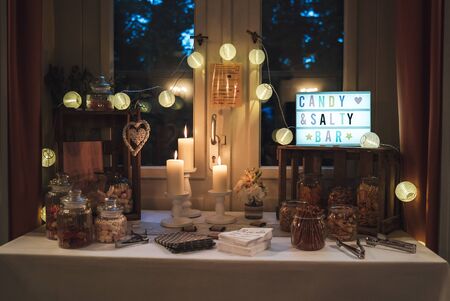 Salty and candy bar of several kinds of nuts, chips, salty snacks and colorful candies in glass jars decorated with candles on white clothed table.の写真素材