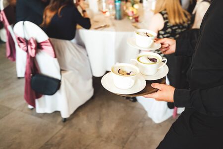 Man in formal clothes carrying three soup mugs of green pea cappuccino soup. Celebration, party,...