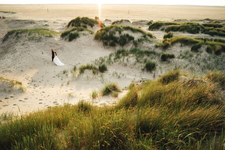 Happy smiling marriage couple walking in sand dunes. Barefoot bride and groom enjoys sand walk in evening sunshine. Wedding day concept.の写真素材
