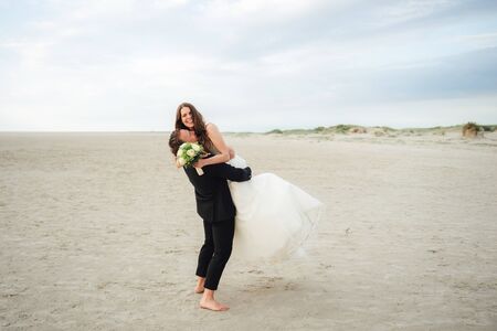 Wedding couple standing and hugging at sandy beach. Groom lifting bride to air. Smiling cheerful bride looking to camera. Wedding day concept.の写真素材