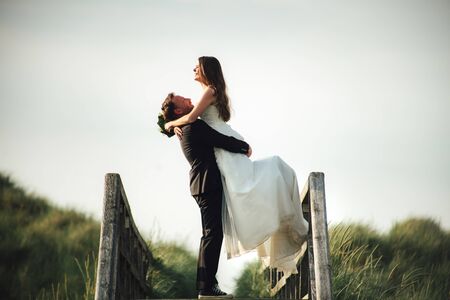 Happy and smiling wedding couple embracing on wooden bridge in evening sun light. Groom lifting bride to the air. Copy space. Wedding day concept.の写真素材