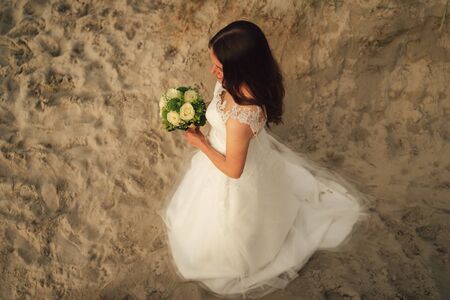 Smiling attractive brunette bride holding wedding bouquet. White roses flowers. Evening light on sandy beach. Wedding day concept.の写真素材