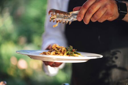 Man serving portion of pasta salad with vegetable and sauce. Male hands holding serving tongs and white plate. Outdoor background.の写真素材