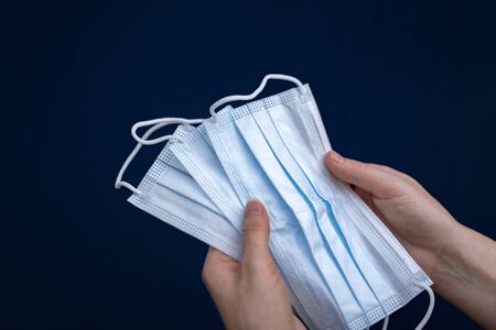 Closeup of female hands holding three surgical disposable face masks isolated on dark blue...