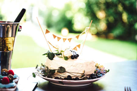 Small white cake decorated with fresh flowers and berries. Outdoor background. Celebration, party, birthday or wedding concept.の写真素材