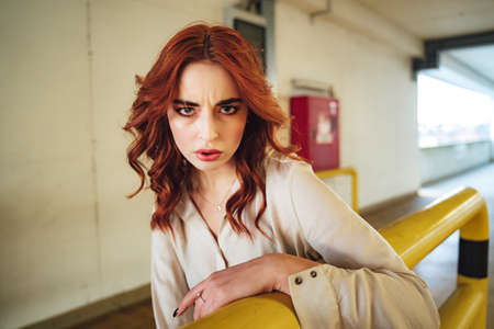 Portrait of frowning young female model leaning on yellow railing. Millennial girl with beautiful long red hair.の写真素材