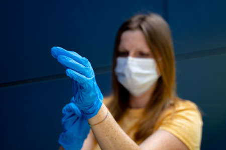 Woman wearing face mask putting on blue protective gloves. Focused on gloves. Corona virus pandemic protection concept.の写真素材