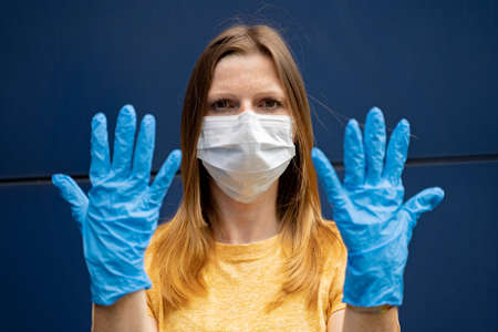Female with medical mask showing hands to camera, wearing blue nitrile gloves over blue background. Corona virus pandemic protection concept.の写真素材