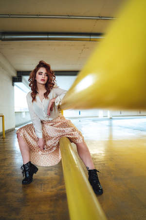 Beautiful caucasian girl sitting on yellow railing and looking to camera. Female red haired model in long skirt.の写真素材