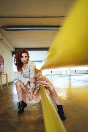 Beautiful caucasian girl sitting on yellow railing and looking to camera. Female red haired model in long skirt.の写真素材