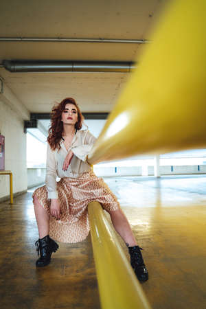 Beautiful caucasian girl sitting on yellow railing and looking to camera. Female red haired model in long skirt.の写真素材