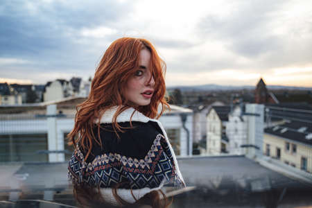 Beautiful millennial girl standing on terrace and looking over shoulder to camera. Attractive female...