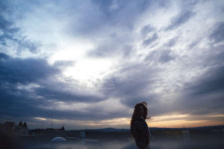 Silhouette of young woman against evening overcast sky. Town buildings in background. Image.の写真素材