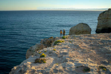 Mid distance view of couple standing on cliff by seascape against sky at sunsetの写真素材