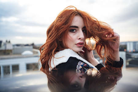 Closeup of beautiful caucasian girl looking to camera and playing with her long wavy red hairs. Female model outdoors, mirror reflection.の写真素材